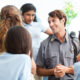 Law enforcement insurance: Diverse group of school children gather around mid adult Caucasian security officer. They are asking him questions about his job. The kids are wearing school uniforms.